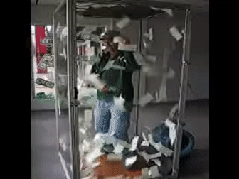 A guy in a glass box grasping at bills flapping around him